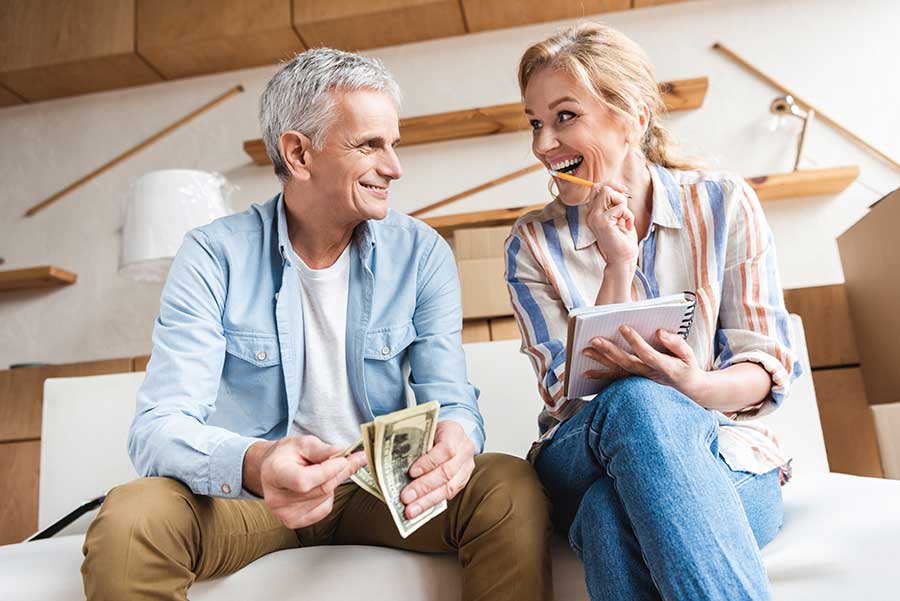 a older couple sitting in a house with moving boxes holding money and a checklist